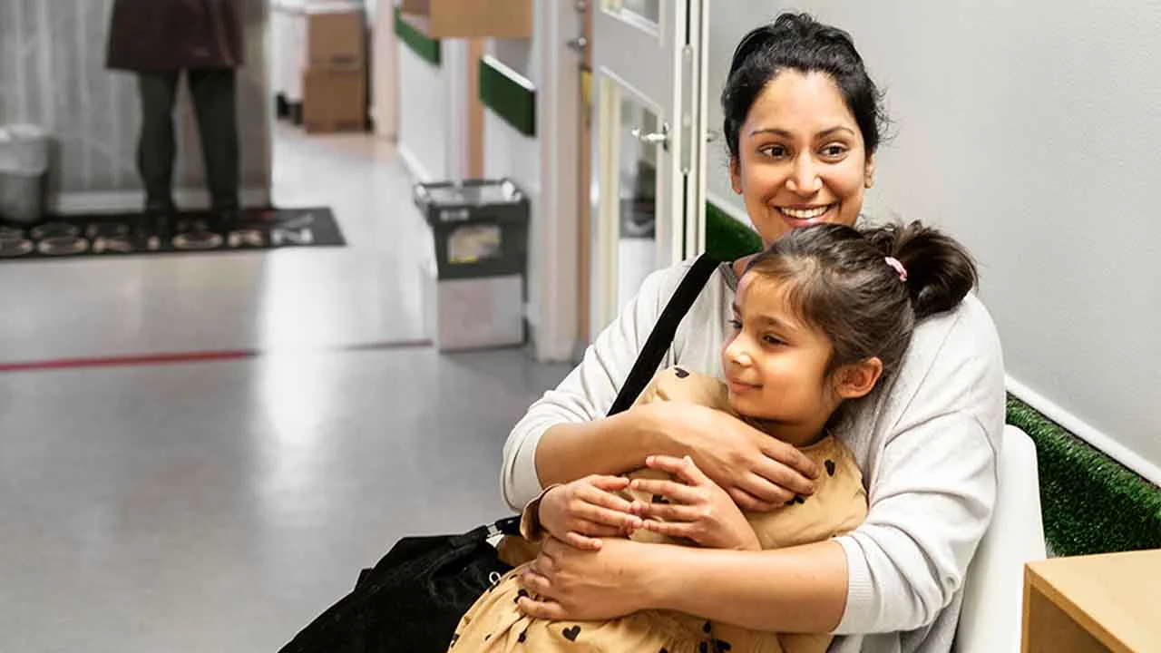 Eine Mutter sitzt auf einem Stuhl im Flur einer Klinik und umarmt ein kleines Mädchen, das auf ihrem Schoß sitzt. 